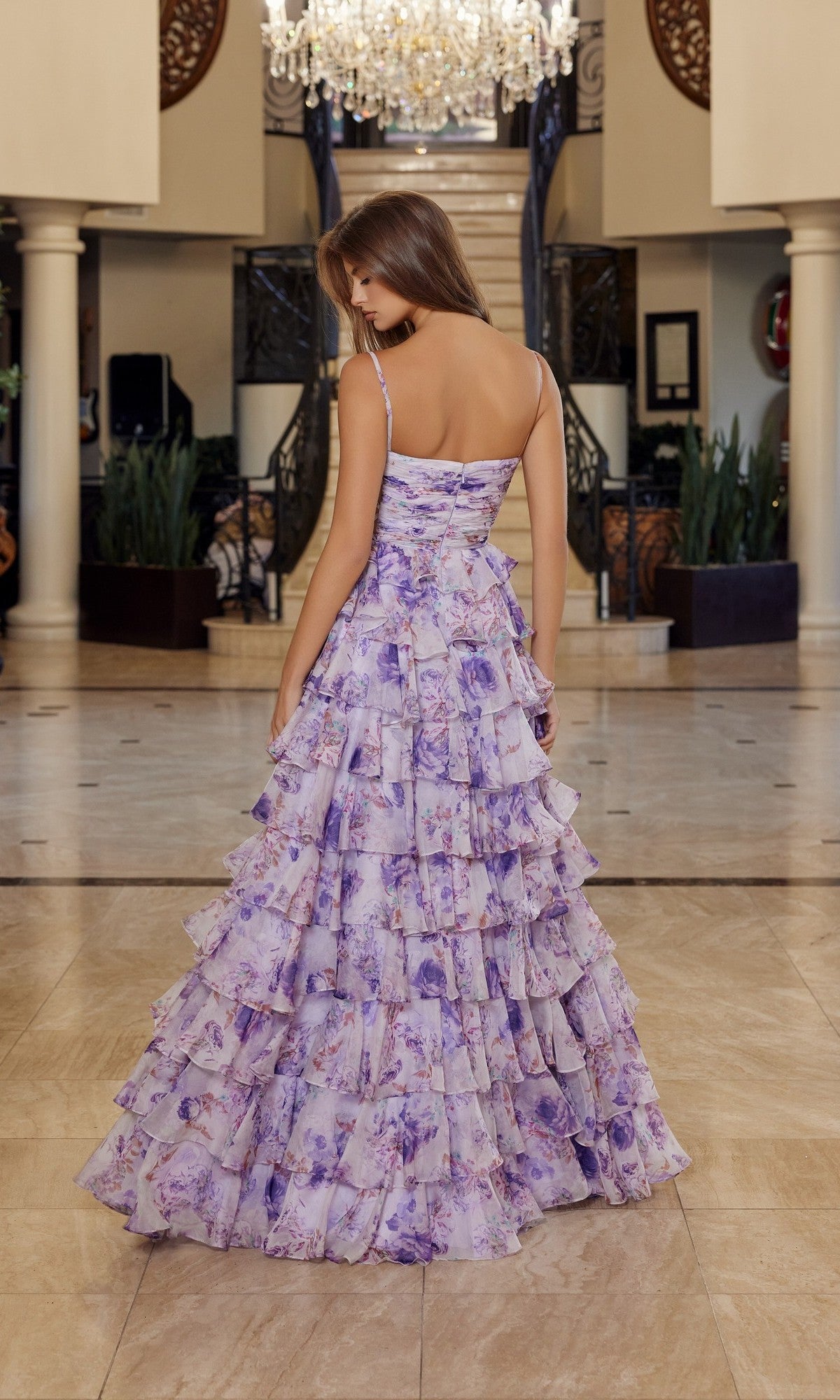 A woman stands indoors facing away, wearing the Nox Anabel Q1777 long lavender floral chiffon prom dress with thin straps and a ruffled A-line skirt. Marble flooring, a staircase, and a chandelier are seen in the background.